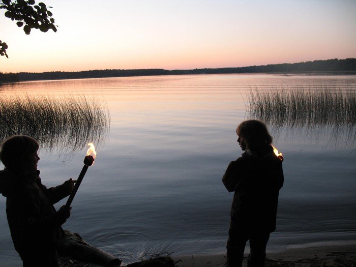 Ein junger Mensch mit einer Fackel, eine weitere Person und ein See in der Abenddämmerung.