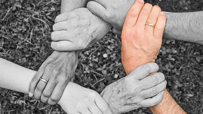 Vier Arme verschlungen in einem Kreis über Gras, eine Hand farbig hervorgehoben.